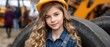 © pvl0707 - Young child with a yellow hard hat smiles while standing near large tires in a busy warehouse filled with workers and machinery during the day