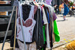 © Adam - A close-up shot of an outdoor clothing rack displaying various garments, including a white embroidered folk corset, a plaid shirt, a leather jacket, and a neon green top, set on a paved area on a sunn