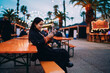 © BullRun - Cheerful mixed race woman browsing on mobile phone while sitting outdoors, happy hispanic girl using smartphone application at evening fairground, authentic digital nomad life, copy space.