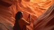 © Envision - Side view of a woman using a smartphone to photograph dramatic orange sandstone walls inside a narrow slot canyon, travel adventure concept with warm evening light