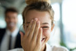 © AI_images - Woman smiling while covering one eye with hand in fun gesture, colleague visible beside her slightly blurred, bright office interior, natural window light