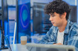 © whyframeshot - focused young Asian male programmer with curly hair is working intently on his laptop in a modern data center office. The background features bright blue neon LED-lit monitor