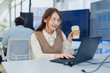 © whyframeshot - professional female software developer with glasses sitting at her desk, holding a coffee cup while intently working on her laptop. The scene captures a modern business environment