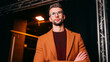 © dinatychynska - Confident male speaker wearing orange coat and glasses standing backstage with arms crossed before presentation, representing public speaking, leadership, business conference and professional training