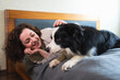 © Юлия Завалишина - Smiling woman playing with cute puppy dog border collie on bed at home indoor. Owner girl stroking holding dog friend sitting on sofa in living room. Love for pets friendship support team concept