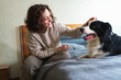 © Юлия Завалишина - Smiling woman playing with cute puppy dog border collie on bed at home indoor. Owner girl stroking holding dog friend sitting on sofa in living room. Love for pets friendship support team concept