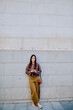 © we.bond.creations - Young woman smiling, standing outdoors by a wall, wearing a brown cardigan and brown trousers, using a mobile phone