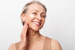 © SHOTPRIME STUDIO - Smiling mature woman with short grey hair applies white face cream on cheek. Natural skincare concept with healthy glowing skin and anti aging beauty routine on white studio background.
