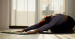 © Binzari - In a living room, a woman practices stretching and pilates on a mat, focusing on her fitness routine as the morning sun filters through the window.