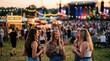 © Mey - A group of young women laugh and chat together at an outdoor music festival. They are standing on a grassy field, surrounded by a large crowd of people enjoying the event.