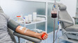 © Esther - Man donating blood, with a full blood bag hanging from an IV stand, in a sterile medical clinic environment.