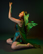 © faestock - portrait of female model wearing fantasy fairy costume, green dress, cloak, butterfly wings and crown. full length sitting pose, kneeling on floor, dark studio background.