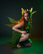 © faestock - portrait of female model wearing fantasy fairy costume, green dress, cloak, butterfly wings and crown. full length sitting pose, kneeling on floor, dark studio background.