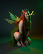 © faestock - portrait of female model wearing fantasy fairy costume, green dress, cloak, butterfly wings and crown. full length sitting pose, kneeling on floor, dark studio background.