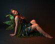© faestock - portrait of female model wearing fantasy fairy costume, green dress, cloak, butterfly wings and crown. full length sitting pose, kneeling on floor, dark studio background.