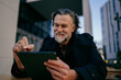 © BullRun - Happy senior professional using digital tablet with stylus, cheerful mature man working on creative project at outdoor cafe, digital nomad lifestyle, authentic representation, career growth.