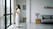 © mashimara - Woman in casual attire holding a mug while looking out a large window