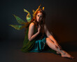 © faestock - portrait of female model wearing fantasy fairy costume, green dress, cloak, butterfly wings and crown. full length sitting pose, kneeling on floor, dark studio background.