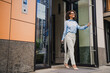 © deagreez - Businesswoman leaves a modern office building smiling while carrying a stylish bag in an urban city scene