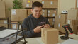 © Krakenimages.com - Man holds smartphone, taps screen and reaches for parcel in building while checking labels; small business focus.