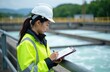 © miss irine - Asian woman engineer wears hard hat and safety vest. She checks clipboard with data at water treatment plant outdoor. Facility monitors water quality.