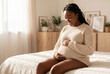 © Meow Creations - Happy pregnant african american woman sitting on bed holding belly in bright bedroom