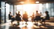 © wiwi - A blurred, warm-toned image depicts a group of silhouetted figures seated around a conference table in a modern office setting, suggesting a business meeting or discussion.