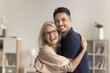 © fizkes - Beautiful mature woman cuddling her young adult son, laughing, pose in domestic room, look for camera with positive, joyful expression. Family attachment, love, support. Happy Mothers Day celebration