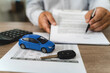 © onephoto - Hands reviewing car loan finance document contract with payment schedule table, blue toy car model and key on paperwork, calculator on desk, representing auto finance agreement analysis, vehicle lease