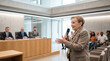 © forenna - Elderly woman with hearing aids speaking confidently at a local council public forum at a municipal hall, microphone in hand, a row of counc 3