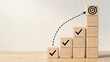 © Jie - Business goal achievement concept wooden blocks tower growing in height with checkmarks and target symbol at top arrow indicating upward trajectory towards success and strategy