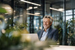 © EricMiguel - Smiling mature businessman in navy dotted blazer and open collar striped shirt, seated at laptop amid lush office plants and glass partitions