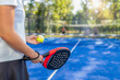 © Microgen - Close-up of padel racket and ball held by player before serve, with blurred doubles match in background on outdoor court, strong sports concept