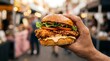 © sbgraphic - A hand holding a delicious fresh burger with lettuce and tomato at a busy street food market