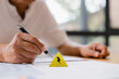 © Teelapon - Close-up of a person holding a pen near a warning sign with lightning symbol on documents, representing electrical risk, safety inspection, and hazard assessment in workplace or business environment.