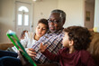 © Marko Geber - Grandfather reading a story to grandchildren on the sofa at home