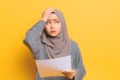 © hilmiyah - Confused young Asian woman wearing hijab is thinking hard while holding tab document.