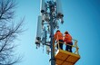 © Viktor - Two workers in orange vests, helmets ascend cell tower on yellow platform lift. Work on antennae, communication equipment against clear blue sky. Technicians install, maintain modern telecom