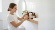 © kodidesign - Smiling woman reaching for skincare bottle in bathroom. Young female with hair in bun using amber glass pump bottles on wooden shelf. Morning self-care routine and beauty products concept