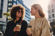 © wolfhound911 - Editorial style gen z women friends chatting happily while walking with takeaway coffee cups in city street, medium close-up urban lifestyle interaction, sunny daylight, modern buildings softly