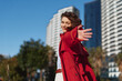 © SHOTPRIME STUDIO - Young woman in a red coat extends her arm toward the camera with a playful smile, standing in a bright urban setting with modern buildings, trees, and blue sky behind.