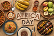 © Muhammad - Africa Day celebration food flat lay, traditional African cuisine with jollof rice and grilled meat, tropical fruits and wooden blocks text