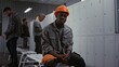 © Framestock - African American Factory Worker Sits on Bench, Wearing Orange Safety Helmet and Grey Jacket, With Metal Lockers and Diverse Colleagues in Background. Industrial Camaraderie and Diversity. Portrait.