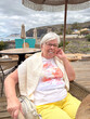© luciano - Smiling Senior Woman Relaxing at Beachside Cafe in Casual Summer Outfit on Overcast Day