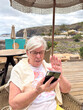 © luciano - Senior Woman Video Calling on Smartphone at Beachside Cafe During Relaxed Coastal Vacation