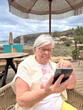 © luciano - Smiling Senior Woman Using Smartphone Outdoors Under Beach Umbrella On Coastal Vacation