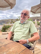 © luciano - Happy Senior Man Relaxing at Beachside Cafe on Vacation, Smiling With Sunglasses and Watch