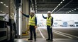 © Liew - Two male workers in reflective vests and hard hats signaling directions inside a modern industrial warehouse with trucks visible
