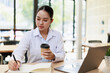 © MINAE - Asian businesswoman freelancer managing her tasks, planning strategies, and writing notes in an agenda while working remotely from a modern office, holding a takeaway coffee