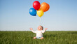 © Alex & M Studio - Happy adorable baby sitting on lush green grass in summer field reaching up to colorful bright balloons against a clear blue sky during a fun outdoor birthday party celebration